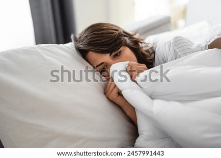 Young sick woman wrapped up in a blanket up to her nose lying in bed. Female person suffering from heat temperature, feeling cold and flu. Exhaustion due to illness. Royalty-Free Stock Photo #2457991443