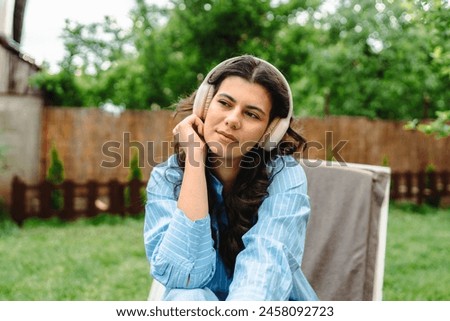 One young caucasian woman is enjoying music on her wireless headphones while laying on easy chair in her backyard
