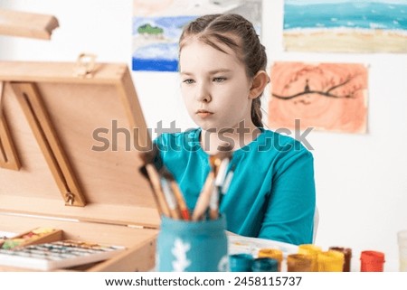 Portrait of a little girl artist, she looks with a concentrated gaze at an easel, there are many paints and brushes on the table. In the background are her drawings. High quality photo