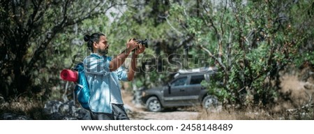 Male photographer with a camera on a hike by car. Young handsome guy with a backpack and a professional camera takes pictures of nature on a hike among the trees in the mountains