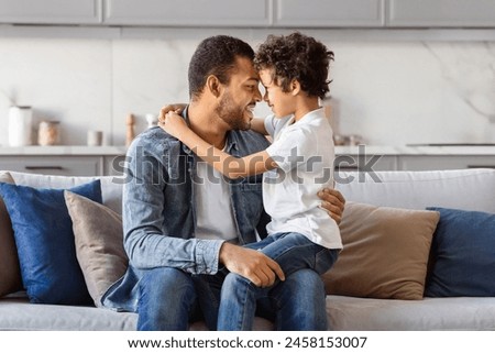 African American man is seated on the armrest of a couch, with one arm wrapped protectively around a little boy who is sitting beside him. They appear to be engaged in a bonding moment. Royalty-Free Stock Photo #2458153007