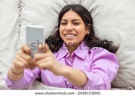 A middle eastern woman is laying comfortably in bed, holding her phone at arms length to take a selfie, looking directly at the phones screen, capturing the moment in a casual and relaxed manner.