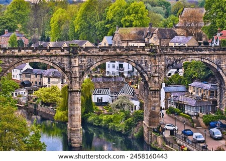 A picturesque view of a stone bridge arching over a river in a quaint village, surrounded by lush greenery and historic buildings. Royalty-Free Stock Photo #2458160443