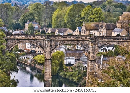 A picturesque view of a stone bridge arching over a river in a quaint village, surrounded by lush greenery and traditional houses. Royalty-Free Stock Photo #2458160755