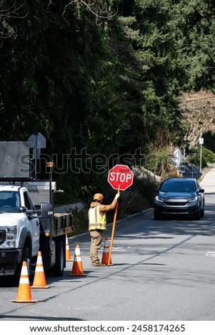City workman with temporary stop sign managing traffic around large work truck in in street with safety cones marking it
