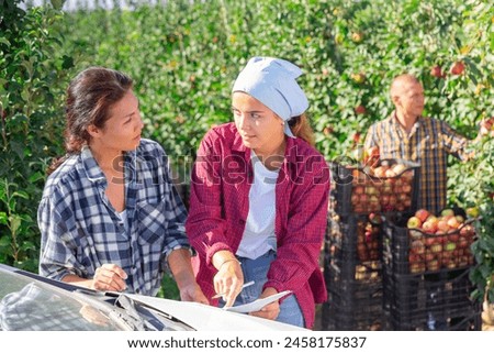 Focused female farmers discussing and signing some papers while standing near car in farm fruit garden during summer harvest of apples