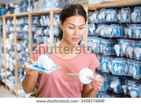 Adult Asian woman choosing bulbs in housewares store. She's holding two packs in hands and comparing them.