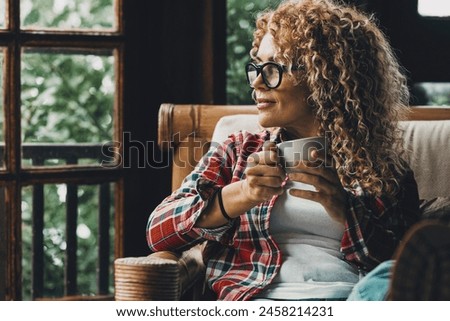 One middle age attractive woman relaxing at home sitting on the chair and looking outside enjoying afternoon time and indoor leisure activity alone. Independent female people indoor. having relax Royalty-Free Stock Photo #2458214231