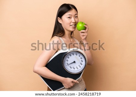 Young Chinese woman isolated on beige background with weighing machine and with an apple