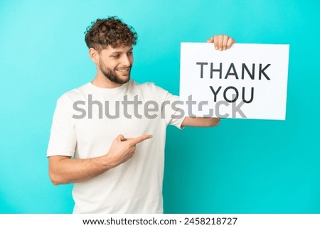 Young handsome caucasian man isolated on blue background holding a placard with text THANK YOU and  pointing it