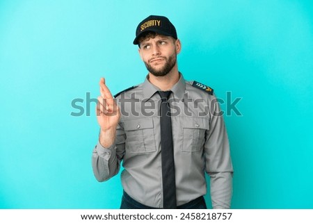 Young security man isolated on blue background with fingers crossing and wishing the best