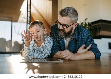 Little boy watching cartoon movie on tablet with father, lying on floor. Dad explaining technology to son, digital literacy for kids.