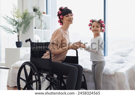 Happy Caucasian woman and daughter doing hair curling. Young mother in wheelchair holds hands of her little daughter on weekend morning in bedroom. Royalty-Free Stock Photo #2458324565