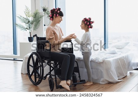 Young mother in wheelchair holds hands of her little daughter on weekend morning in bedroom. Happy Caucasian woman and daughter doing hair curling. Royalty-Free Stock Photo #2458324567