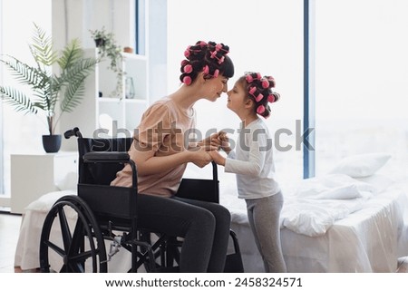 Happy Caucasian woman and daughter doing hair curling. Young mother in wheelchair holds hands of her little daughter on weekend morning in bedroom. Royalty-Free Stock Photo #2458324571