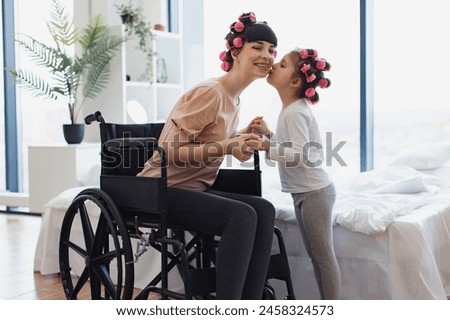 Happy Caucasian woman and daughter doing hair curling. Young mother in wheelchair holds hands of her little daughter on weekend morning in bedroom. Royalty-Free Stock Photo #2458324573