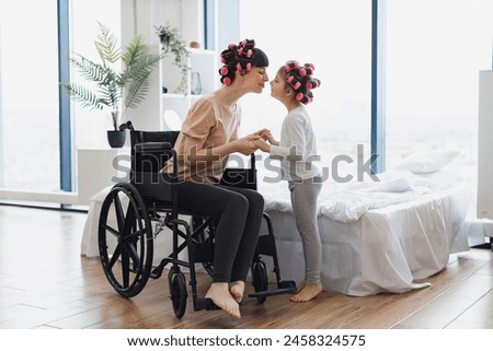 Young mother in wheelchair holds hands of her little daughter on weekend morning in bedroom. Happy Caucasian woman and daughter doing hair curling. Royalty-Free Stock Photo #2458324575