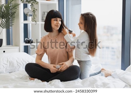 Little cute girl in casual outfit brushing her beautiful mothers hair sitting near big panoramic window in bedroom. Happy Caucasian family having fun on bad on weekend morning. Royalty-Free Stock Photo #2458330399