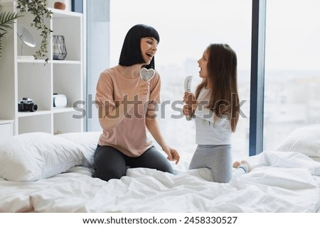 Happy Caucasian family having fun on a weekend morning. Young mother sings with her little adorable daughter using combs instead of microphones while sitting on bed. Royalty-Free Stock Photo #2458330527