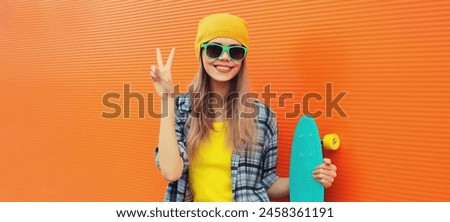 Summer portrait of happy cheerful stylish young woman with skateboard in colorful clothes, yellow hat posing on orange background on city street