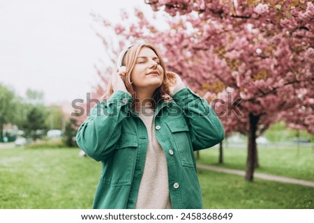 Portrait of charming 30s woman in massive headphones listening to music in the park. Music lover enjoying music. Spring background with a branch of blooming sakura. Positive emotion concept