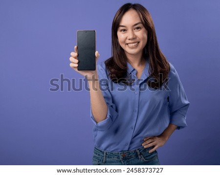 Smiling Asian woman in blue shirt pointing to her smartphone, showcasing the screen, isolated on a purple background