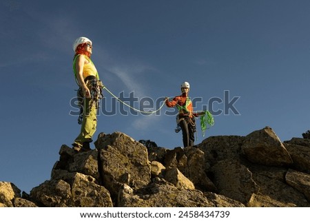 Two people are climbing a mountain, one of them wearing a green harness. The other person is wearing a red jacket