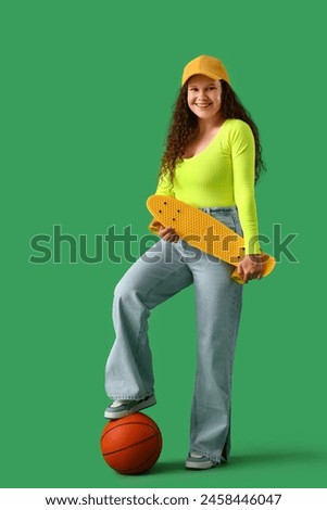 Beautiful young stylish woman with skateboard and ball on green background