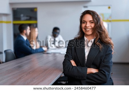 Confident young businesswoman stands in modern office, smiling. Colleagues discuss in background. Pro at work in formal attire, successful female leader, team meeting, workplace dynamics. Royalty-Free Stock Photo #2458460477
