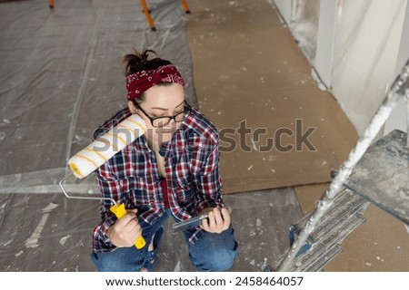A woman with glasses and a roller in her hands is watching a video tutorial on painting walls. High quality photo