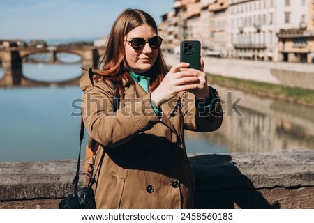Attractive young female tourist is exploring new city. Redhead girl holding a smartphone on famous Old bridge in Florence. Traveling. Happy optimistic girl walking in city and makes selfie