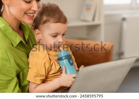 A woman is sitting while holding a child in her arms, multitasking by using a laptop. Mother and son watching cartoon together, home interior, copy space