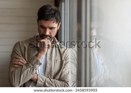 A man stands in front of a window, deep in thought, with his hand resting on his chin. The natural light illuminates his profile as he gazes outside, deep in contemplation. Royalty-Free Stock Photo #2458595903