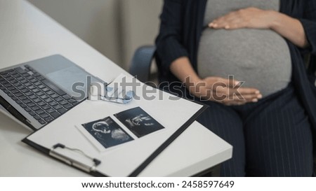 A pregnant woman works on a laptop in the office and looks at a photo from an ultrasound scan of the fetus. Belly close-up.