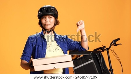 Close up shot of part time courier holding takeaway pizza boxes, showing thumbs down sign. Food delivery cyclist doing disapproving finger shaking gesture, saying no, isolated over studio background