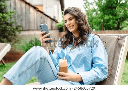 One young caucasian girl is relaxing on easy chair while drinking coffee and using mobile phone in her backyard

