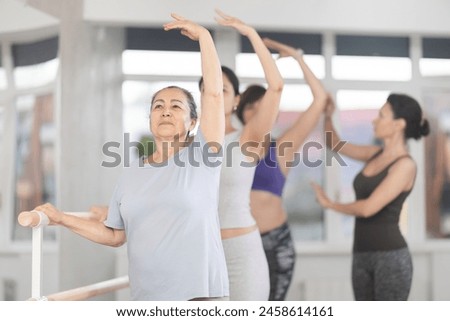 Focused elderly woman learning ballet technique at beginners class with group of female enthusiasts of different ages under guidance of instructor, practicing third position at barre in light studio Royalty-Free Stock Photo #2458614161