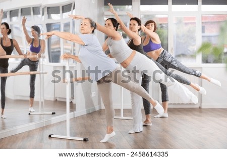 Active elderly woman finding joy and maintaining health by practicing ballet with female group in beginner class, learning graceful arabesque at barre in choreography studio Royalty-Free Stock Photo #2458614335