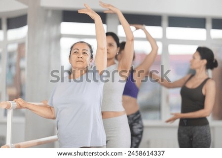 Focused elderly woman learning ballet technique at beginners class with group of female enthusiasts of different ages under guidance of instructor, practicing third position at barre in light studio Royalty-Free Stock Photo #2458614337