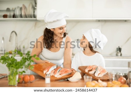 Cheerful middle-aged woman holding wooden board with fish pieces standing at kitchen-table with little daughter