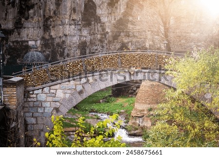 a sunlit morning scene features a charming stone bridge arching gracefully over a placid stream framed by lush foliage against a rugged rock backdrop. Tbilisi Old Town Royalty-Free Stock Photo #2458675661