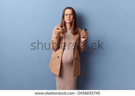 Unhappy dissatisfied young adult pregnant woman wearing dress and jacket holding mobile phone and paper cup with coffee looking at camera with unhappy face posing isolated over blue background