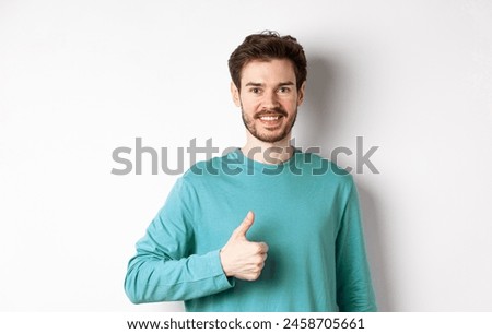 Smiling male model in casual shirt showing thumb up, approve and recommend product, praise excellent choice, standing on white background.
