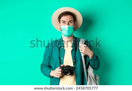  travelling concept. Young guy tourist in medical mask and summer hat travel abroad during coronavirus pandemic, taking pictures on vacation, blue background.
