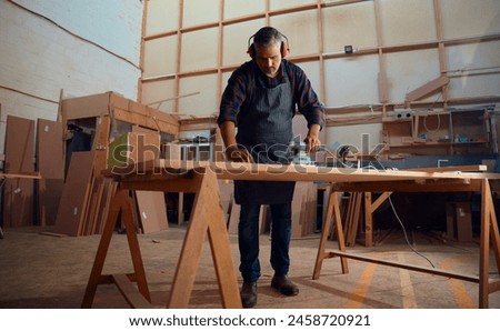 Mid adult man wearing apron and ear muffs and using power tool on wood in woodworking factory