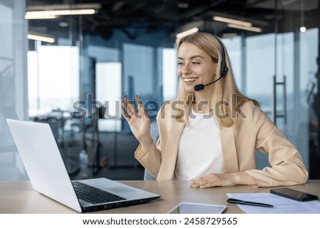 A cheerful professional woman engaging in a video call while working on her laptop in a brightly lit, contemporary office space. Royalty-Free Stock Photo #2458729565