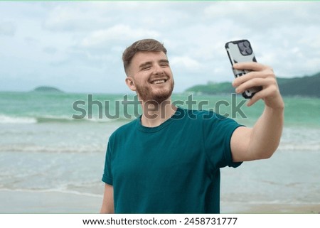 Young guy, handsome happy man at sea is taking picture of hisself, selfie at camera of his phone, using smartphone for social media at summer beach in tropical exotic country. Blogger, vlog