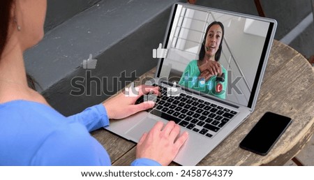 Image of media icons over caucasian woman having laptop image call with biracial colleague. Global business and digital interface concept digitally generated image.