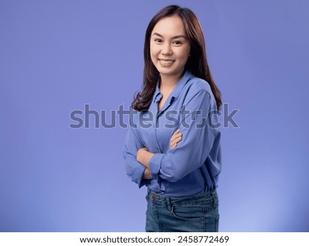 Confident Asian woman in a blue blouse with arms crossed, smiling warmly against a soft purple background