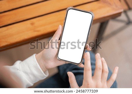 A close-up image of a woman in casual clothes using her smartphone at a table indoors. A white-screen smartphone mockup for displaying graphic ads. people and wireless technology concepts